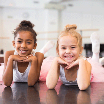 Zwei kleine Ballerinas liegen in Rosa Tütüs auf dem Bauch und lächeln in die Kamera.