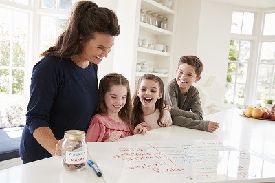 Mutter steht mit ihren drei Kindern an einem Tisch. Sie schauen fröhlich auf eine erstellte Übersicht. Auf dem Tisch steht außerdem ein Glas mit der Aufschrift "Taschengeld".
