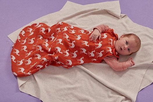 Baby trägt einen rot-gemusterten Musselin-Schlafsack ohne Ärmel und darunter ein geringeltes Langarm-Shirt