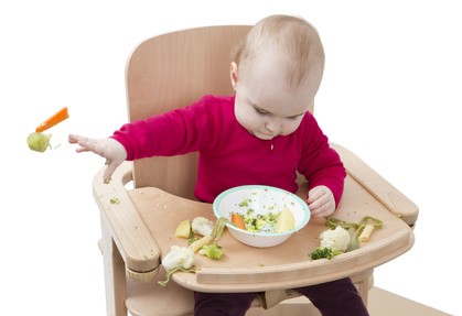 Baby sitzt in einem Hochstuhl und wirft mit Brokkoli, Möhren, Blumenkohl und Kartoffeln, die in der Schale vor ihm waren