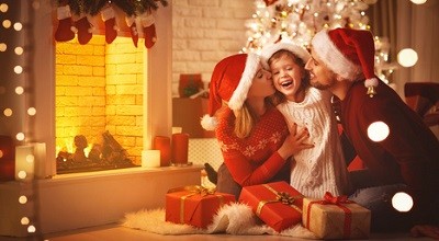 Mutter, Vater und Tochter sitzen eng beieinander vorm Kamin in einem weihnachtlich dekorierten Zimmer. Vor ihnen liegen Geschenke.