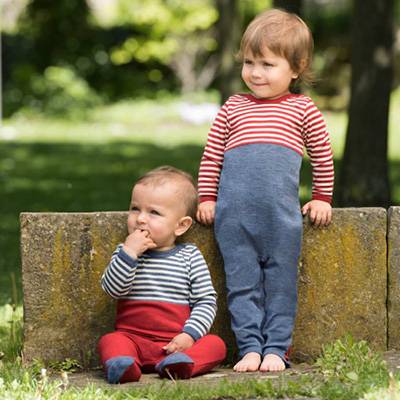 Zwei Geschwister draußen im Garten: Das Baby sitzt mit dem Rücken an eine kleine Mauer gelehnt. Seine große Schwester steht daneben und stützt sich auf der Mauer ab.