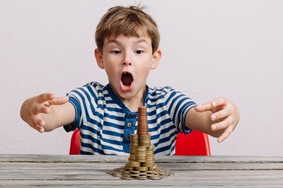 Junge sitzt staunend an einem Tisch. Er hat aus Geldmünzen einen großen Turm gebaut.