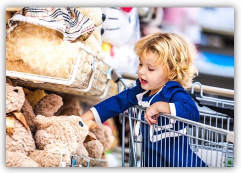 Kind sitzt in einem Einkaufswagen und greift nach einem Teddybär, der im Regal liegt.