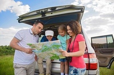 Papa, Mama und zwei Kinder stehen am offenen Kofferraum ihres Autos und schauen auf eine Landkarte.