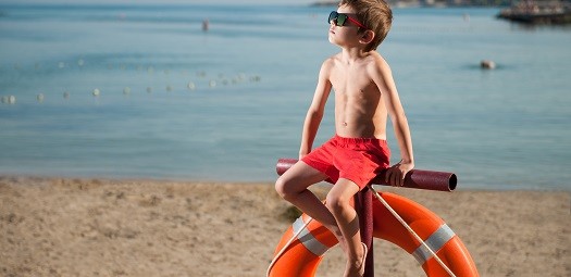 Junge posiert in roter Badehose und mit Sonnenbrille als Rettungsschwimmer. Dabei sitzt er auf einer Halterung eines Rettungsrings und im Hintergrund ist ein See zu sehen.