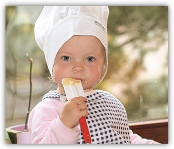 Kind mit Kochmütze auf dem Kopf leckt den Spatel ab, an dem Teigreste kleben