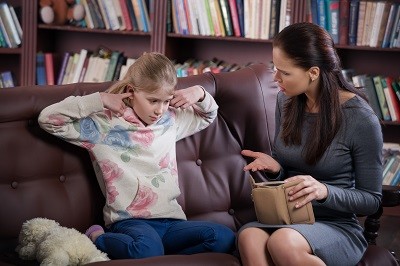 Mädchen sitzt auf dem Sofa und hält sich die Ohren zu. Die Mutter sitzt neben ihr mit einem leeren Portemonnaie und redet auf sie ein.