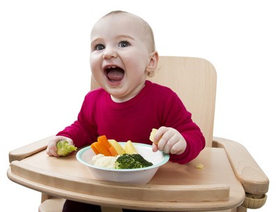 Baby sitzt auf einem Babystuhl, vor ihm steht eine Schüssel mit Möhren, Brokkoli, Kartoffeln und Blumenkohl.