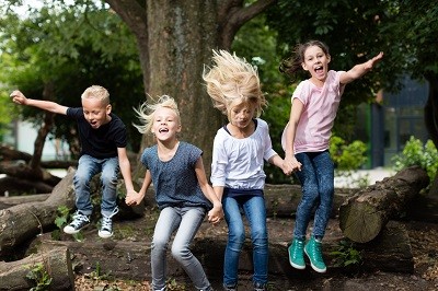 Vier Kinder springen von einem liegenden Baumstamm und halten sich dabei an den Händen.