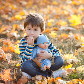 Ein Junge sitzt im Herbst auf einer mit Laub bedeckten Wiese, hält eine Baby-Puppe auf seinem Schoß und isst etwas.