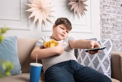 Übergewichtiger Junge sitzt mit einer Schüssel Chips im Arm  und einem Softdrink auf dem Sofa und zappt durchs Programm.