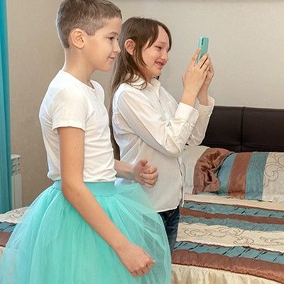 Ein Junge und ein Mädchen stehen nebeneinander vor einem Spiegel. Der Junge trägt ein türkises Tütü und das Mädchen eine Jeans mit Hemd. Sie macht ein Foto von den beiden im Spiegel.