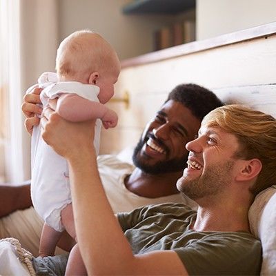 Zwei Männer liegen im Bett und lachen ihr Baby an, das einer von ihnen hochhält. Einer der beiden Männer ist eine Person of Color.