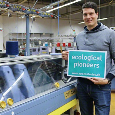 greenstories-Gründer Denver Mielke in den Produktionshallen von disana mit einem Schild auf dem steht "ecological pioneers".