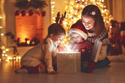 Mutter und ihre zwei Kinder sitzen auf dem Boden in einem weihnachtlich dekorierten Zimmer und schauen in eine glitzernde Box, aus der es leuchtet.