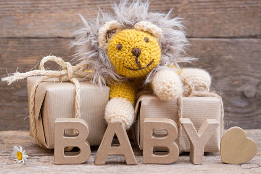 Zwei nachhaltig eingepackte Geschenke liegen auf einer Holzbank. Auf ihnen sitzt ein Kuscheltier-Löwe. Davor steht in Pappbuchstaben das Wort "BABY". Ein Gänseblümchen und ein Deko-Herz aus Holz liegen daneben.