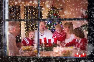 Durchs Fenster sieht man eine am Tisch zusammensitzende Familie. Ein Adventskranz steht vor ihnen, im Hintergrund ein geschmückter Weihnachtsbaum.