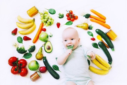 Baby liegt auf dem Rücken und hat ein Salatblatt im Mund. Um es herum liegt verschiedenes Obst & Gemüse.