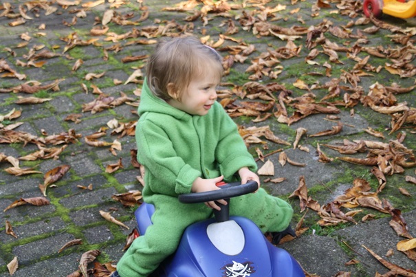 Mädchen sitzt im Overall auf einem blauen Rutschauto und fährt durch das herumliegende Herbstlaub