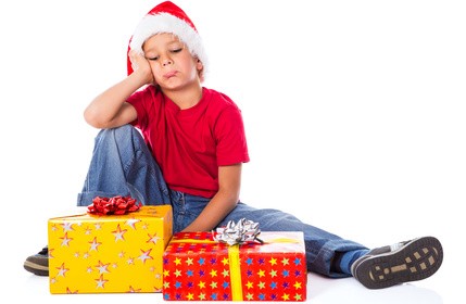 Junge mit Weihnachtsmütze sitzt traurig vor zwei eingepackten Weihnachtsgeschenken.